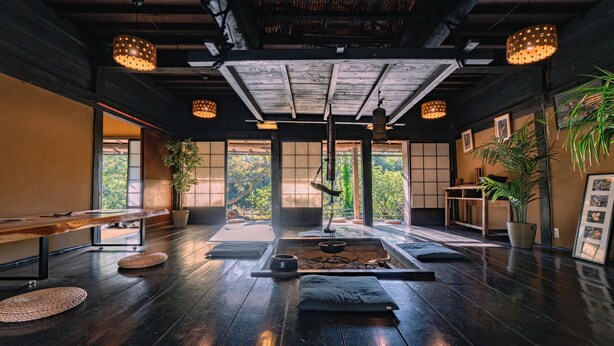 Una habitación amplia con piso de madera, una mesita baja, cojines, una lámpara colgante y vistas al verdor exterior.