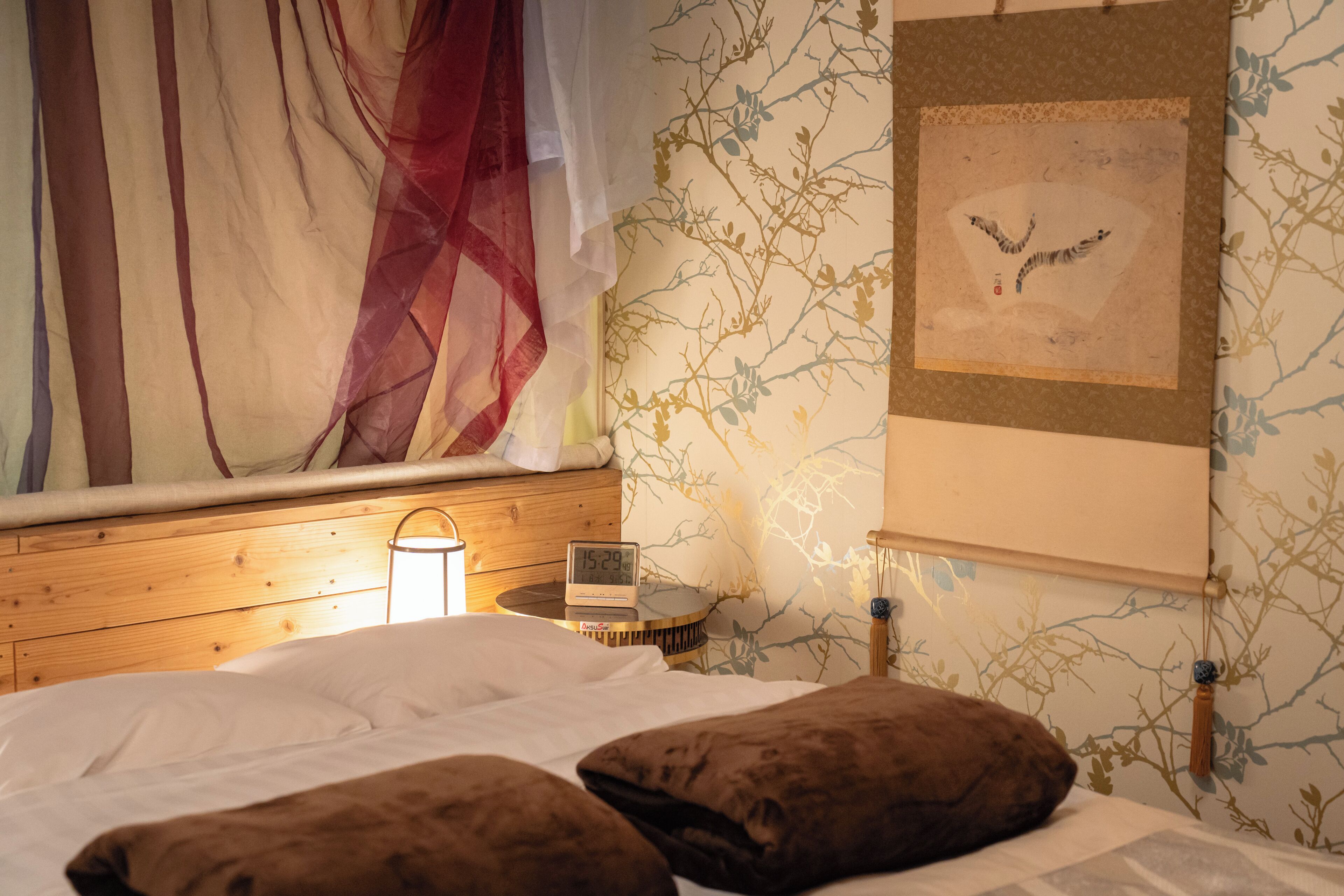 Une chambre à coucher avec un lit, des oreillers, une table de chevet avec une horloge, un tableau accroché au mur et une fenêtre avec des rideaux légers.