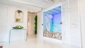 Una zona de recepción con un mostrador de vidrio, un espejo con un arreglo floral, un cartel en la pared y una pared decorativa con una escena de playa.