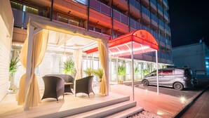 Entrada de un hotel con un área de estar al aire libre techada, un coche aparcado fuera y un edificio de varios pisos.