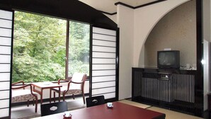 A room with a dining table, chairs, a TV, and a view of greenery through large windows. - Oshuku Onsen Hotel Uguisu (Shizukuishi)