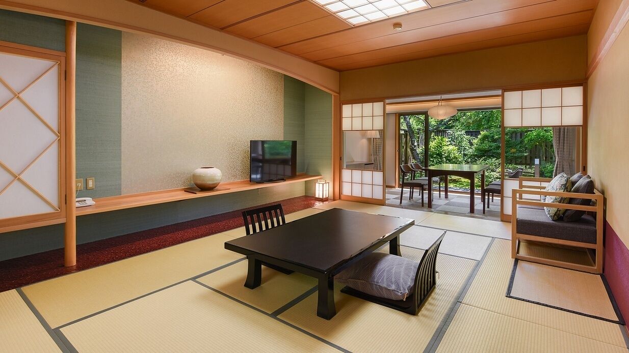 Um quarto japonês tradicional com tatames, uma mesa baixa com almofadas, uma televisão numa prateleira e vista para o verde lá fora.