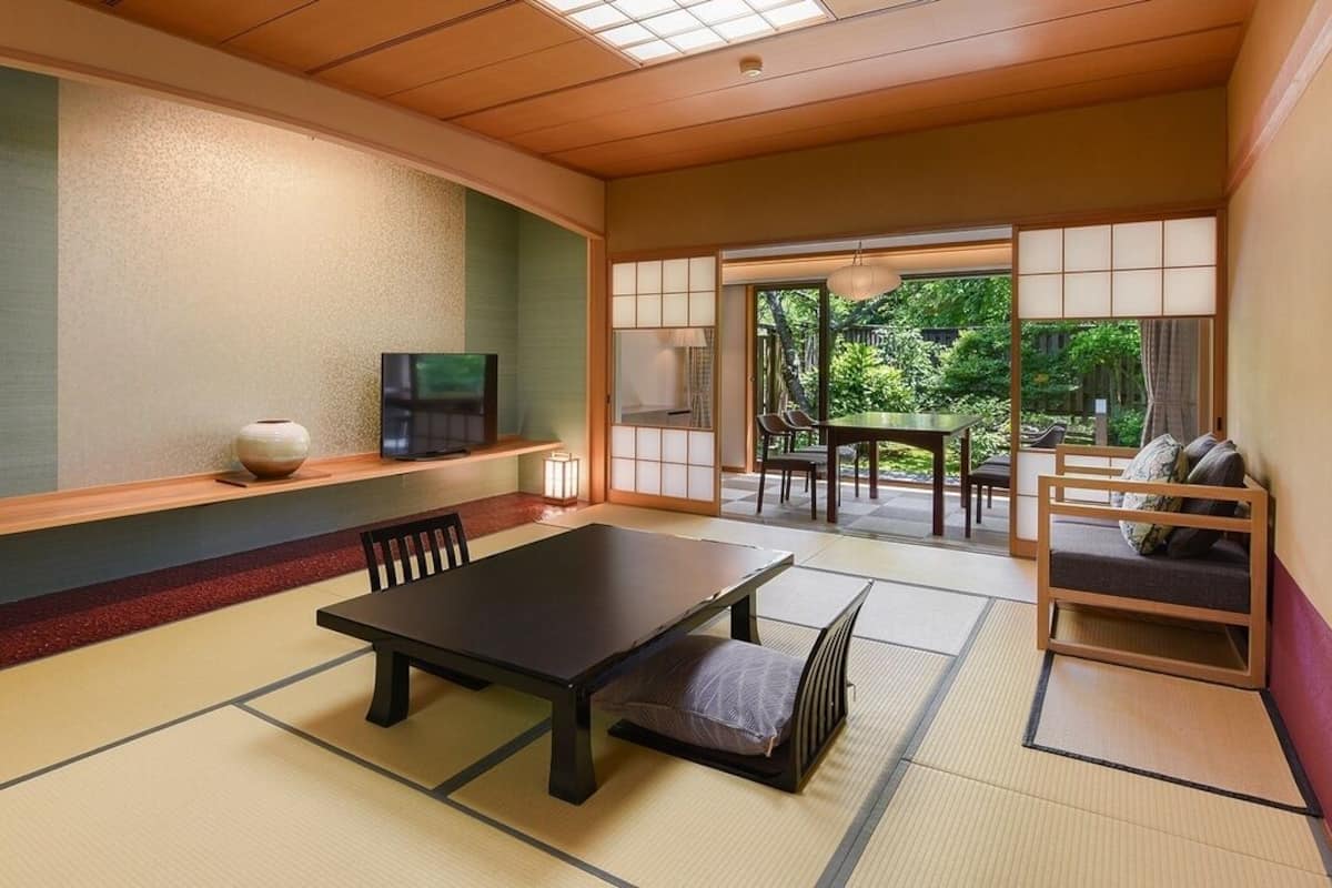 Een traditionele Japanse kamer met tatami vloerbedekking, een lage tafel met kussens, een televisie op een plank en uitzicht op groen buiten.