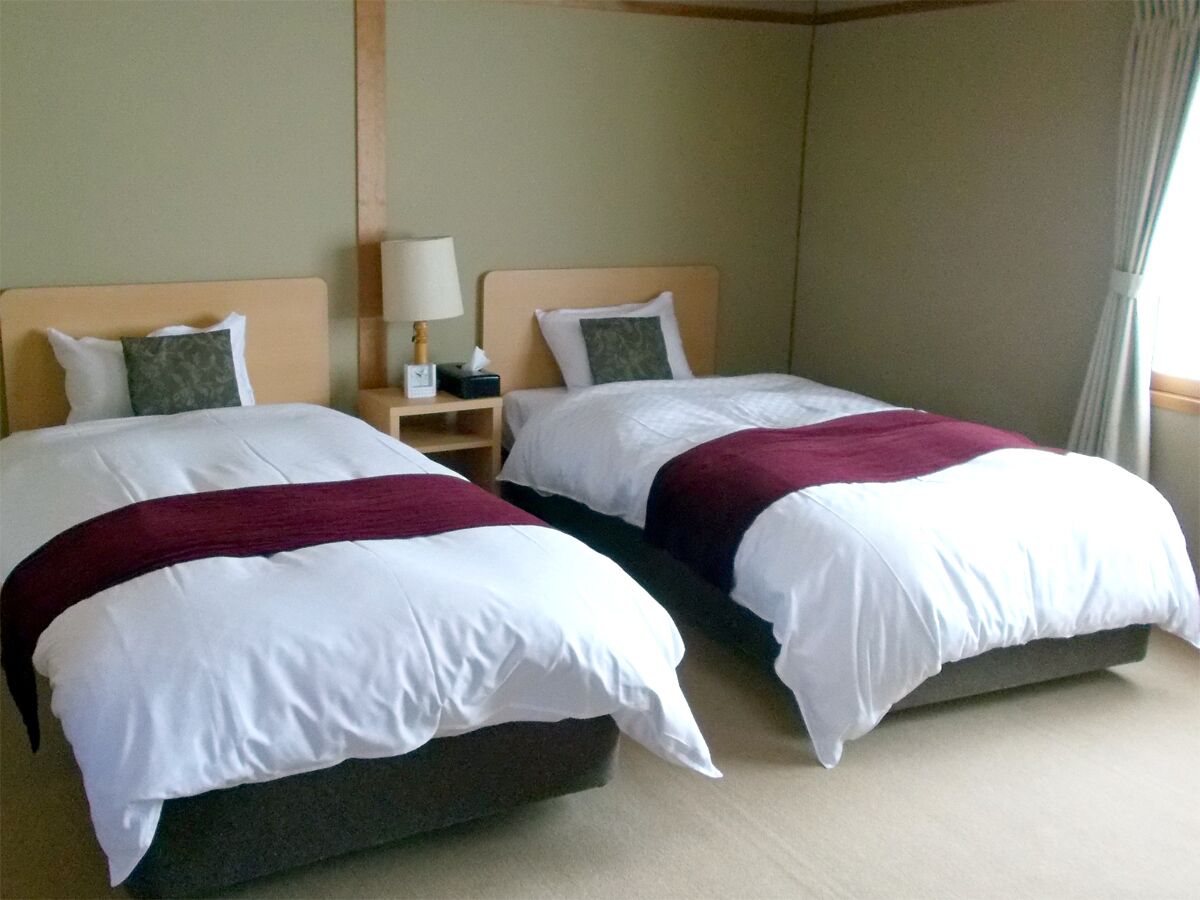 Quarto de hotel com duas camas, cada uma com roupa de cama branca e um cobertor borgonha. Há uma mesa de cabeceira com abajur entre as camas.
