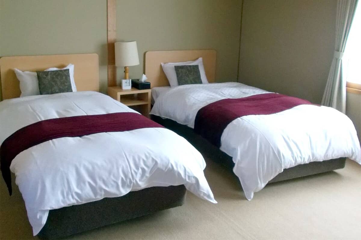 Een hotelkamer met twee bedden, elk voorzien van wit beddegoed en een bordeauxrode plaid. Daartussen staat een nachtkastje met een lamp.