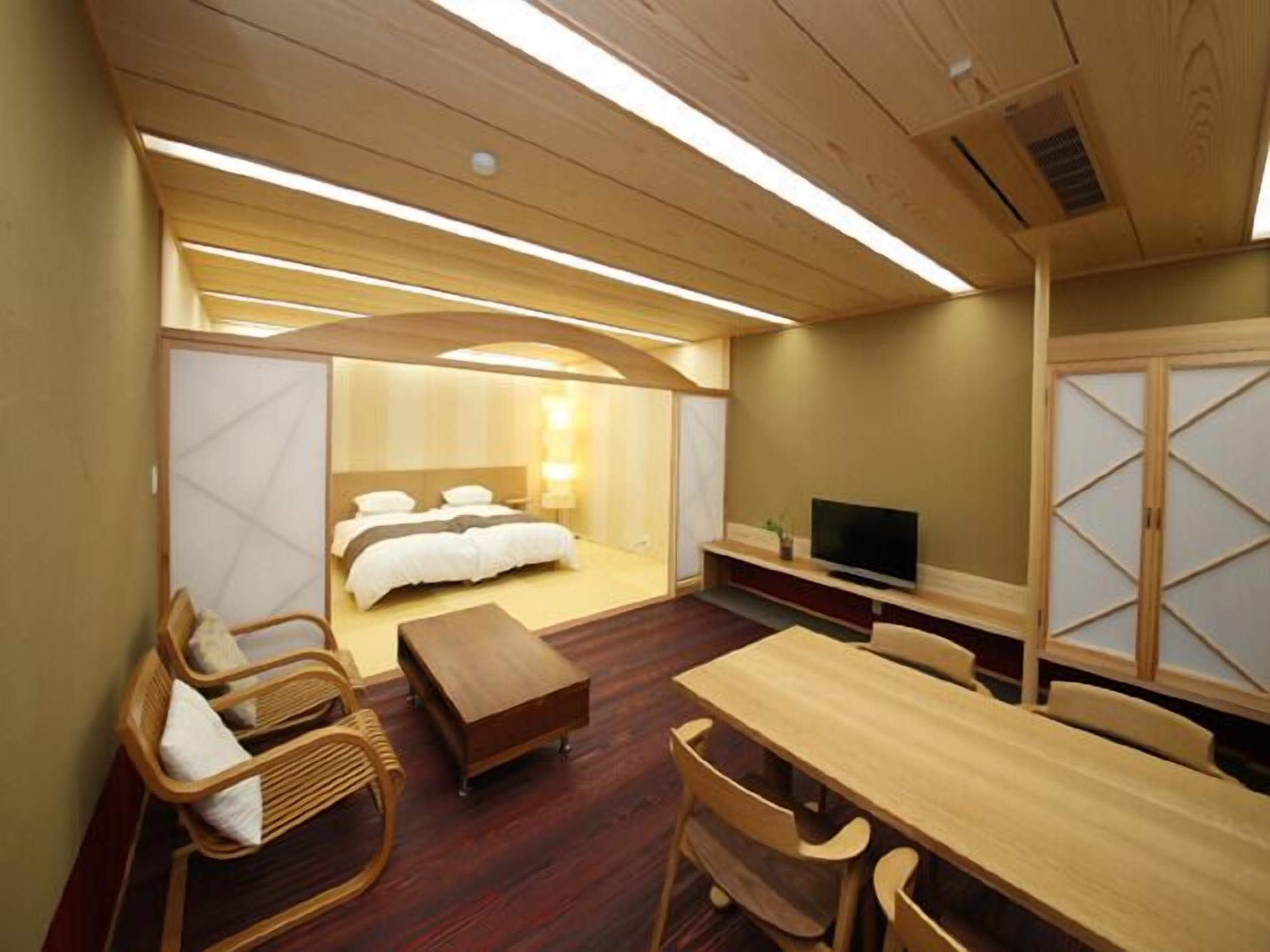 Quarto de hotel com cama, televisão, mesa de jantar de madeira e área de estar com cadeiras.