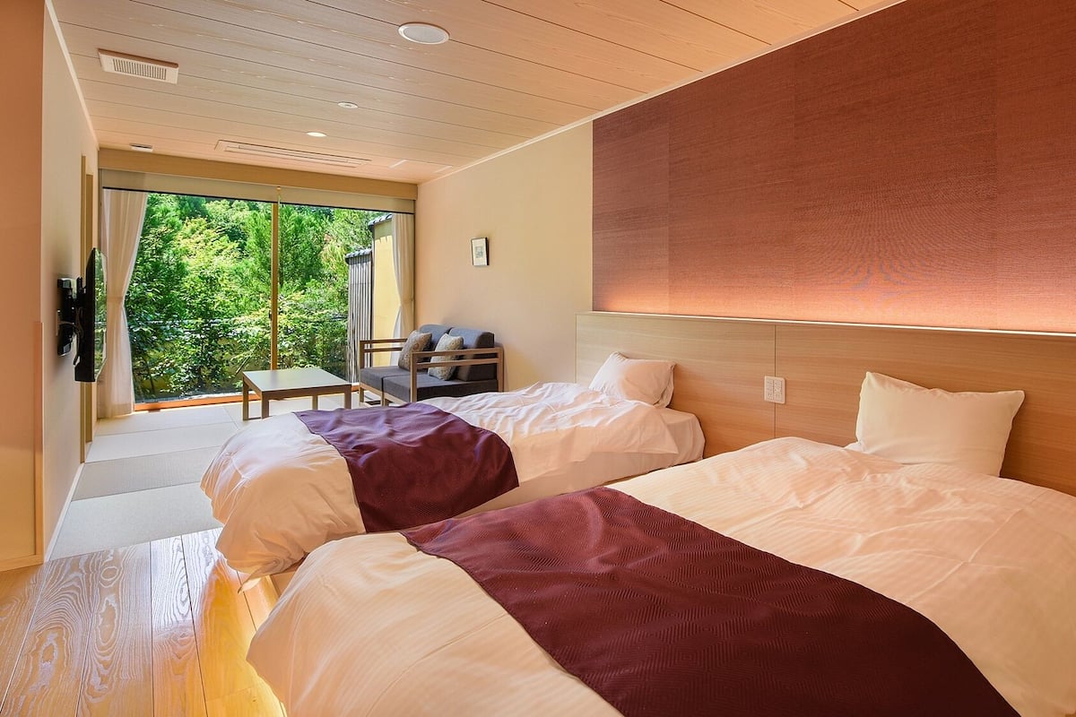 A hotel room with two beds, a sofa, a table, and a view of greenery outside.