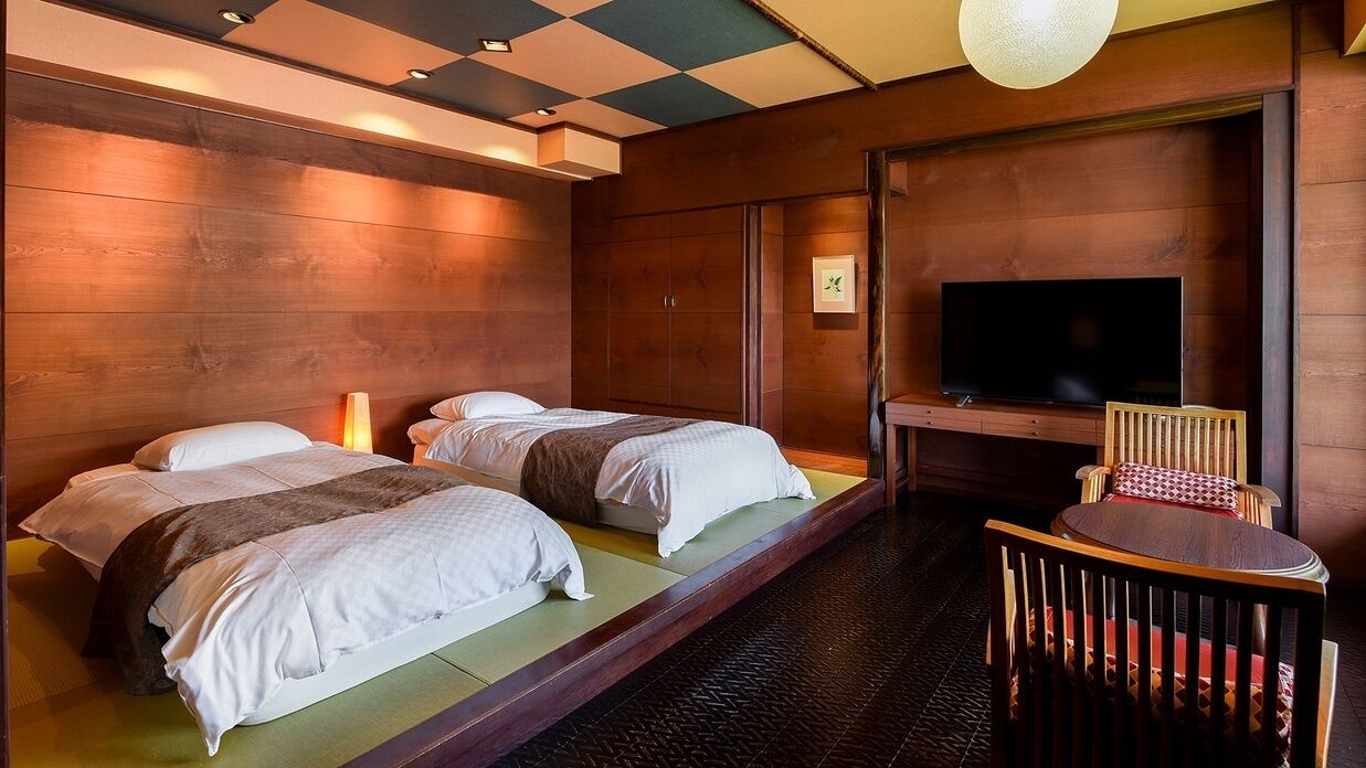 Quarto de hotel com duas camas, uma televisão, uma cadeira de madeira e uma pequena mesa.