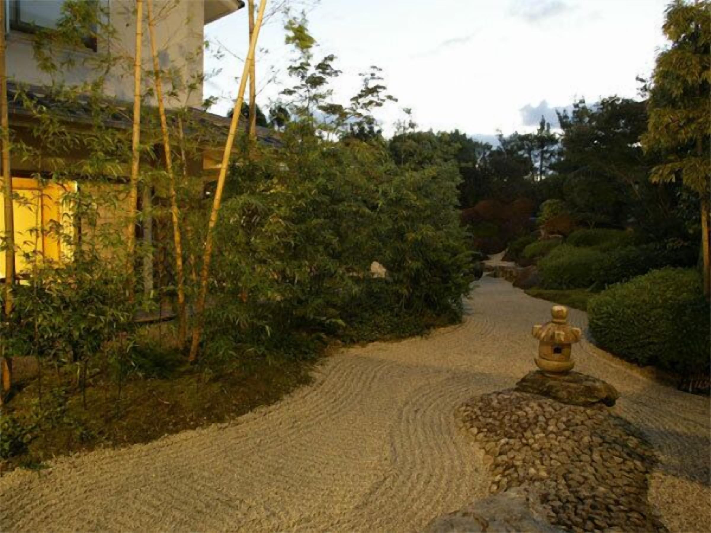Uma trilha de cascalho serpenteia por um jardim com bambu e arbustos diversos.
