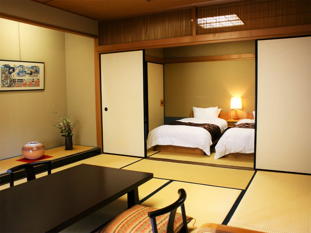 Ein traditioneller japanischer Raum mit Tatami-Böden, einem Bett mit weißer Bettwäsche, einem Tisch mit Stühlen und einer Vase mit Blumen.