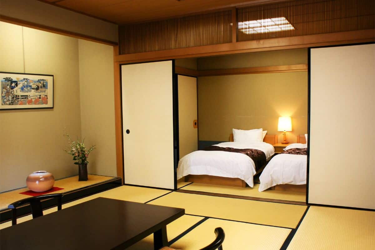 Een traditionele Japanse kamer met tatami vloerbedekking, een bed met wit beddengoed, een tafel met stoelen, en een vaas met bloemen.