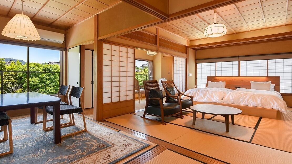 Quarto tradicional em estilo japonês, com tatames, cama baixa, mesa de jantar com cadeiras e vista para o verde lá fora.