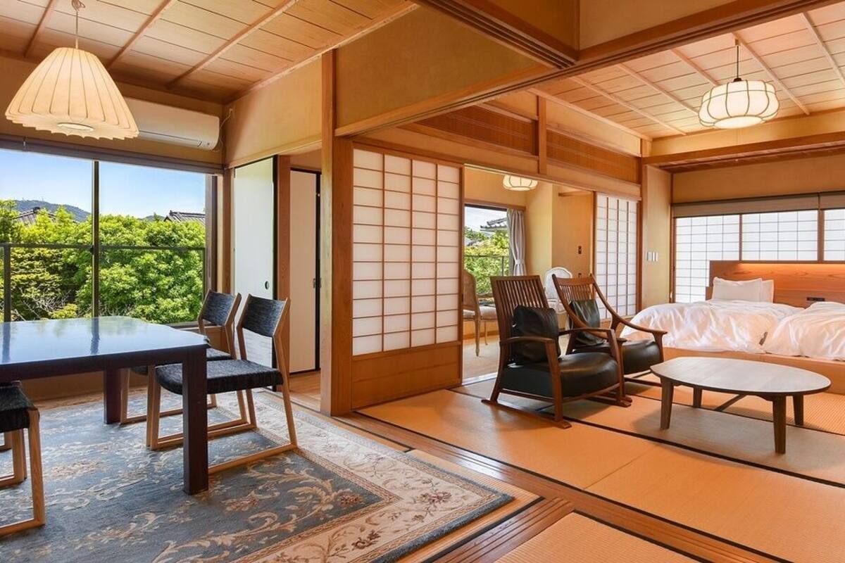 Een traditionele Japanse kamer met tatami vloeren, een laag bed, een eettafel met stoelen en uitzicht op groen buiten.