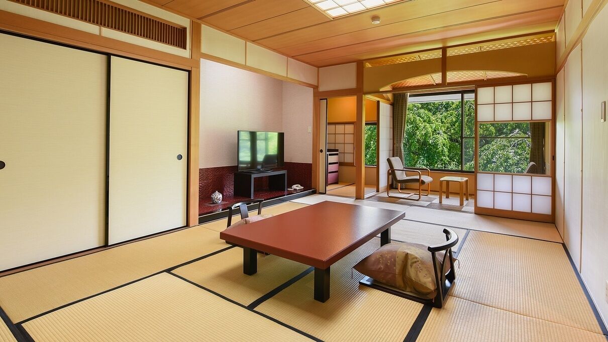 Ein traditioneller japanischer Raum mit Tatami-Böden, einem niedrigen Tisch, einem Fernseher und Schiebetüren, die auf einen Balkon mit Blick auf Bäume führen.