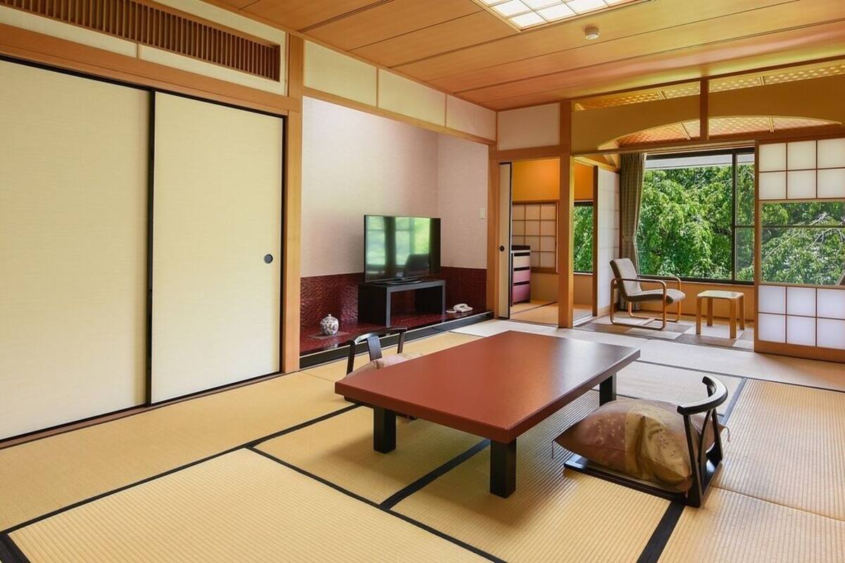 Een traditionele Japanse kamer met tatami vloeren, een lage tafel, een televisie en schuifdeuren die leiden naar een balkon met uitzicht op bomen.