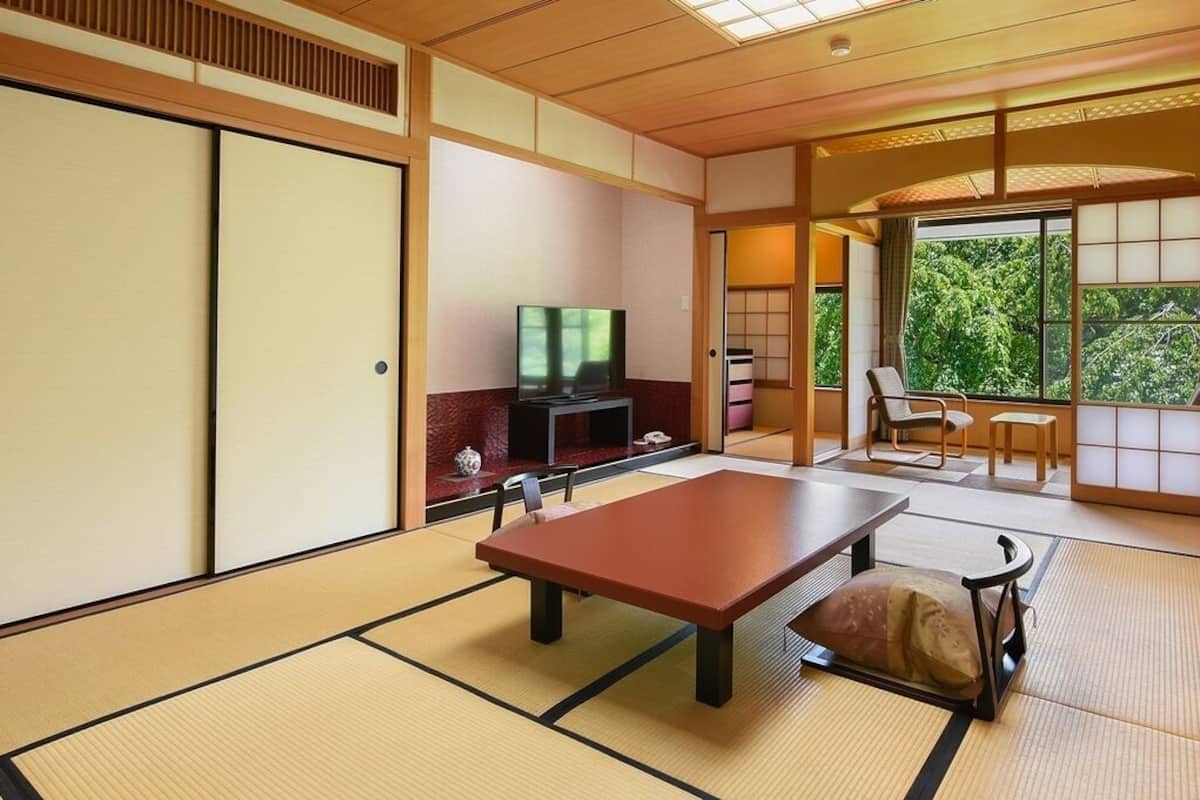 Ein traditioneller japanischer Raum mit Tatami-Böden, einem niedrigen Tisch, einem Fernseher und Schiebetüren, die auf einen Balkon mit Blick auf Bäume führen.