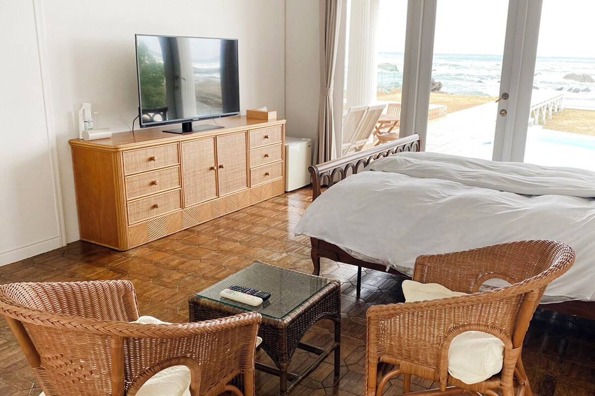 Une chambre d’hôtel avec un lit, des fauteuils en osier, une télévision, un bureau et une vue sur la plage.