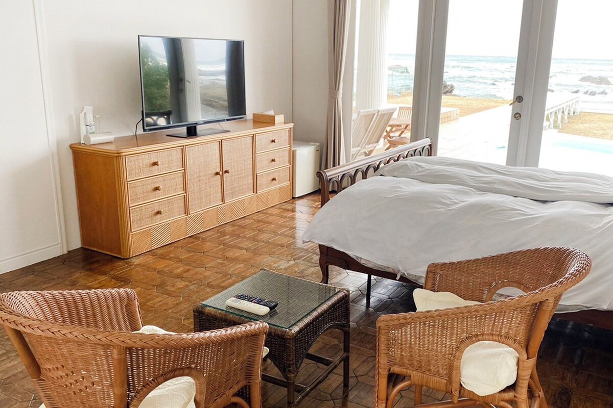 Une chambre d’hôtel avec un lit, des fauteuils en osier, une télévision, un bureau et une vue sur la plage.