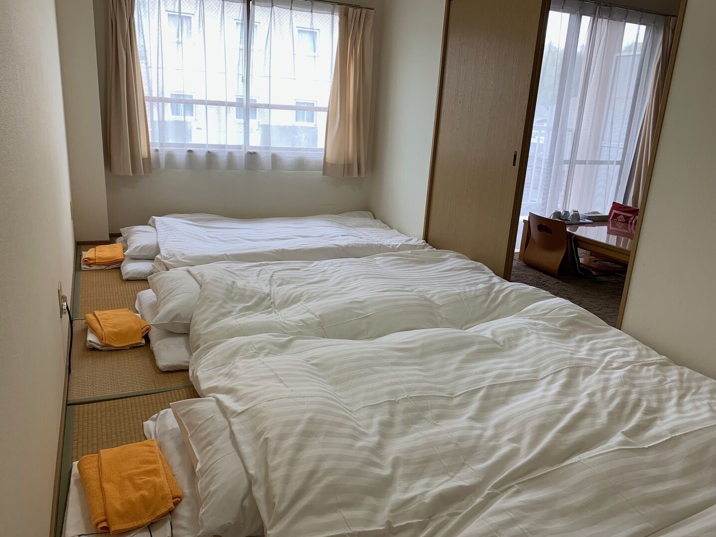 Habitación de hotel con tres camas, una ventana con cortinas, una mesita con una silla y una puerta.