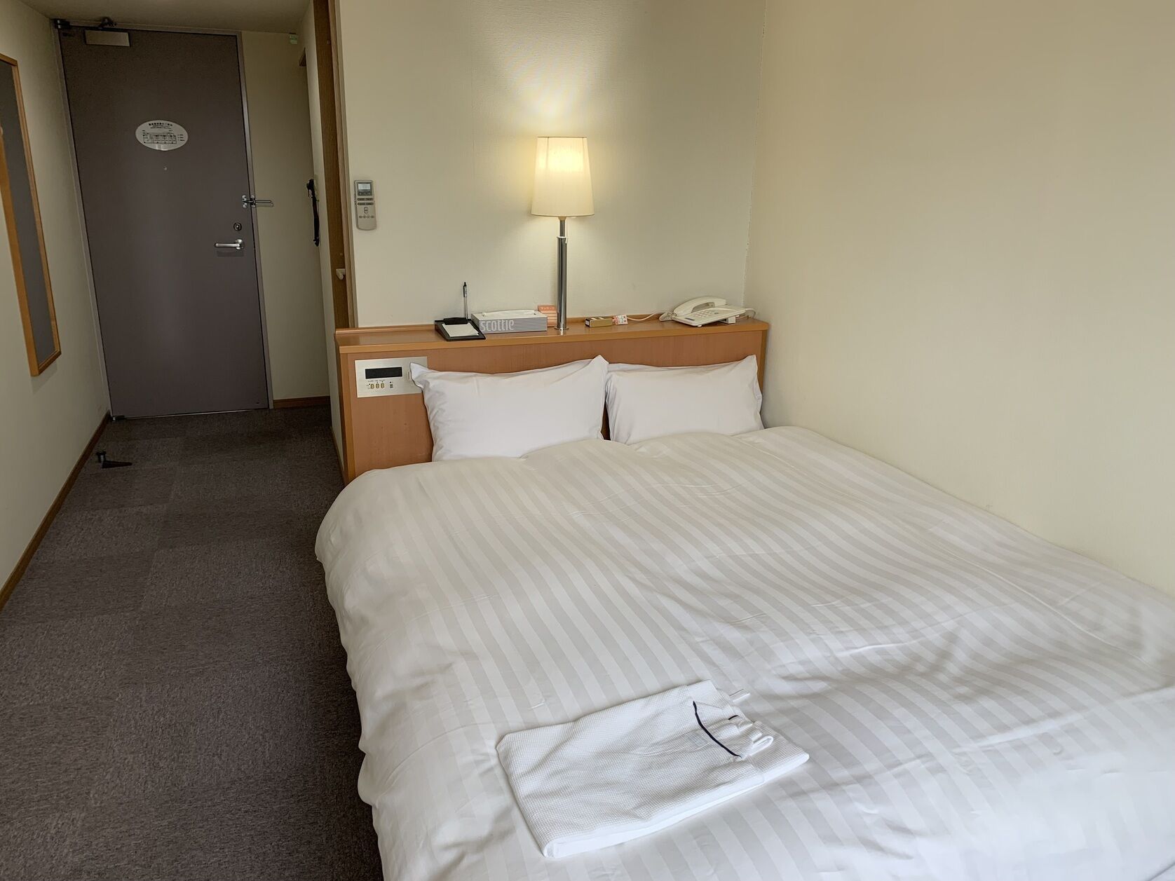 Habitación de hotel con una cama, una lámpara de noche, un teléfono y una puerta.
