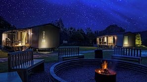 Een sterrenhemel met twee verlichte hutten, buiten zitplaatsen en een vuurkuil in het midden.