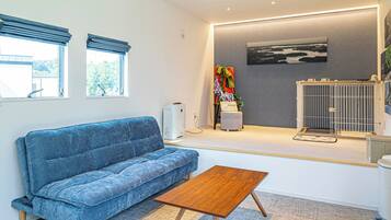 Una sala moderna con un sofá azul, una mesa de centro de madera, un purificador de aire blanco, una pared gris con un cuadro enmarcado y una jaula blanca para mascotas.