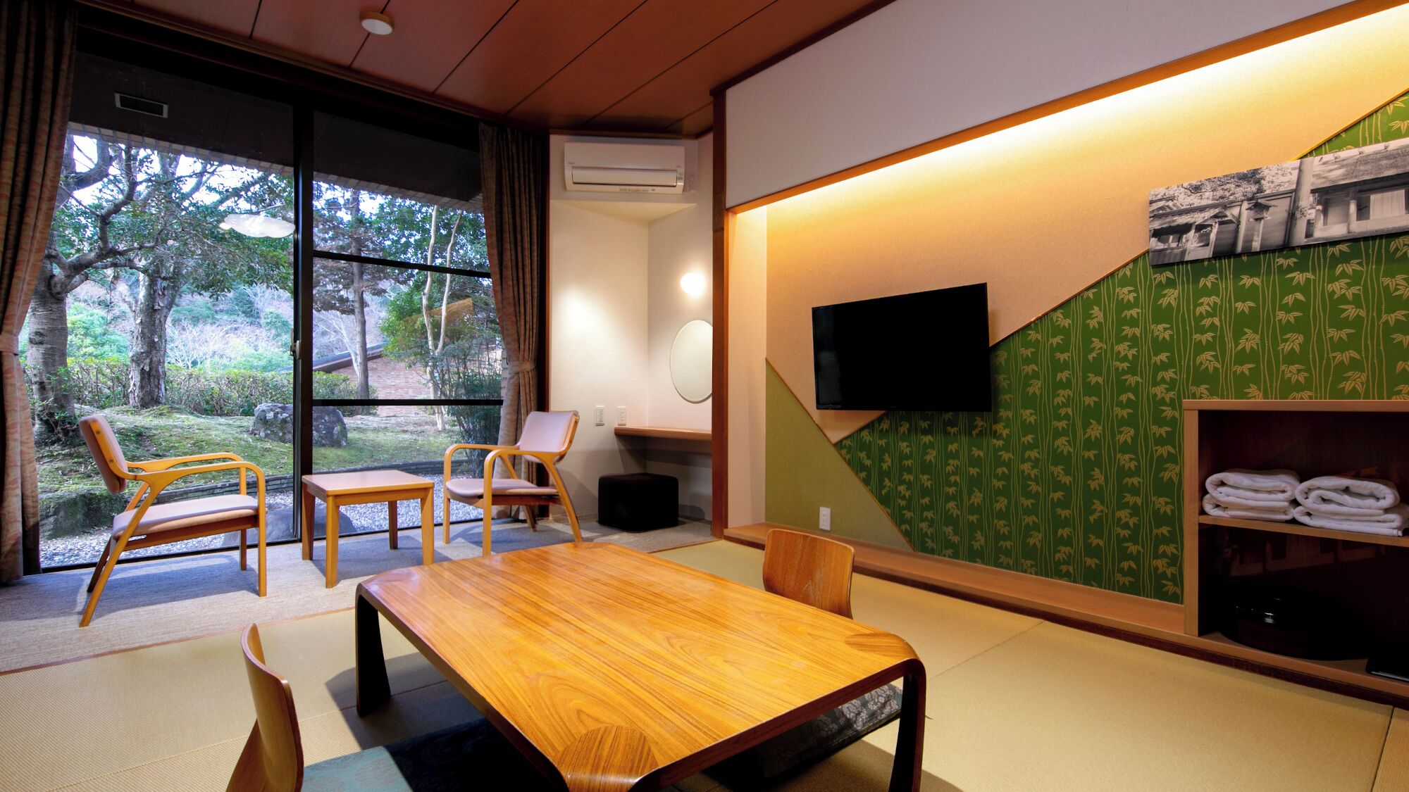 A room with a wooden table, chairs, a TV, and a view of trees through large windows.