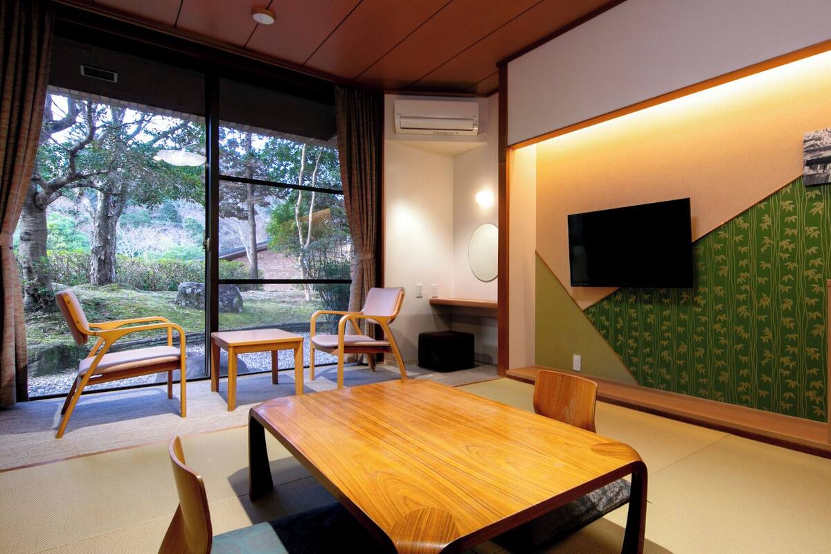 Una habitación con una mesa de madera, sillas, un televisor y vistas a árboles a través de amplios ventanales.