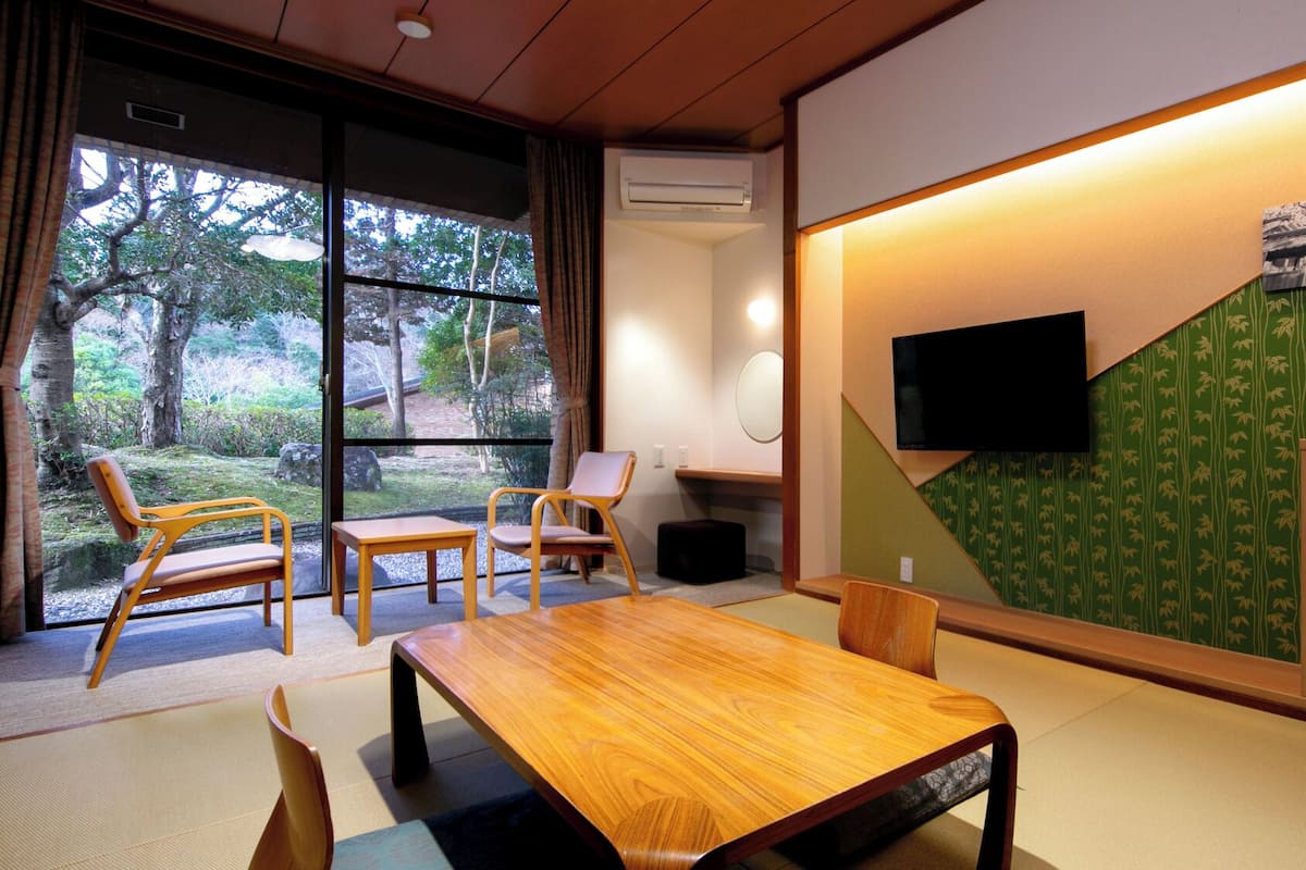 A room with a wooden table, chairs, a TV, and a view of trees through large windows.