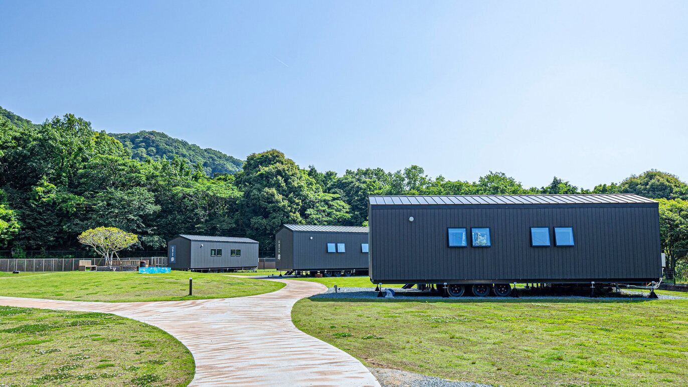 A series of modular cabins with a curved pathway leading to them, set in a grassy area with a forested hill in the background.