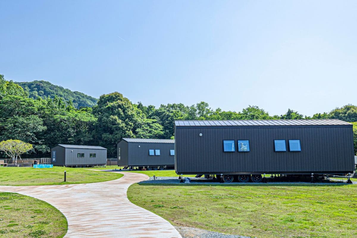 A series of modular cabins with a curved pathway leading to them, set in a grassy area with a forested hill in the background.