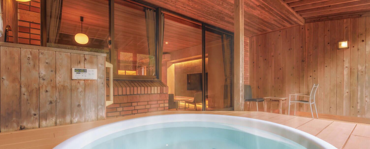 A wooden interior with a hot tub, a brick wall, and a glass door leading to a room with a television.