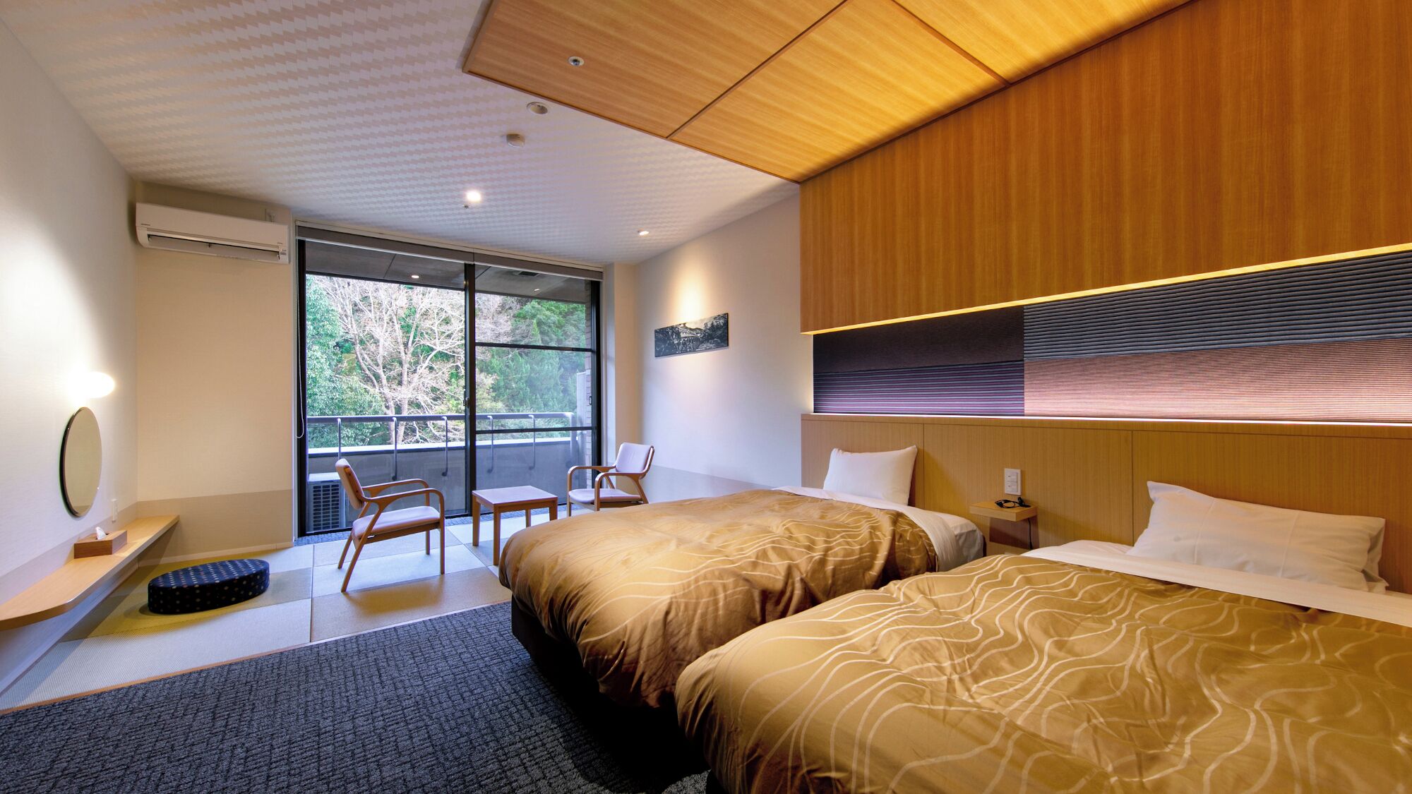 A hotel room with two beds, a desk, a chair, and a large window with a view of trees.
