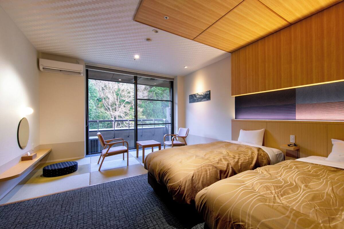 Habitación de hotel con dos camas, un escritorio, una silla y un ventanal con vistas a árboles.