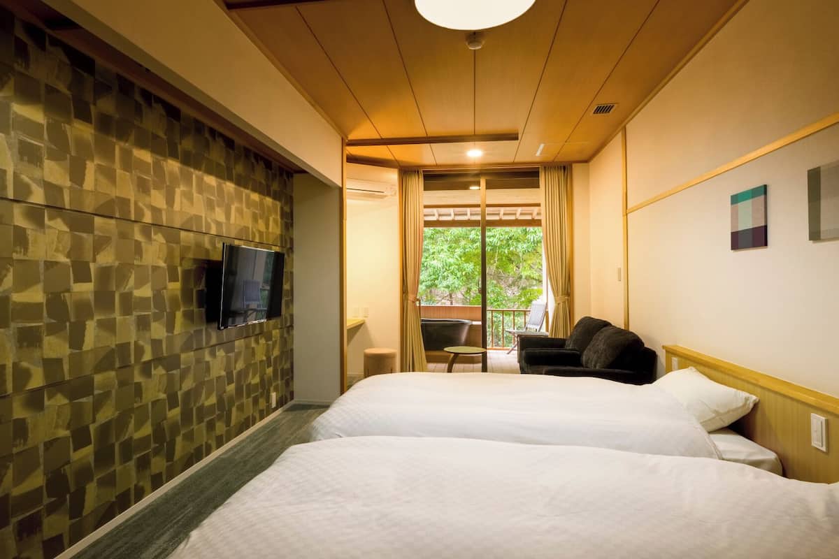 A hotel room with two beds, a TV mounted on the wall, and a view of greenery outside.