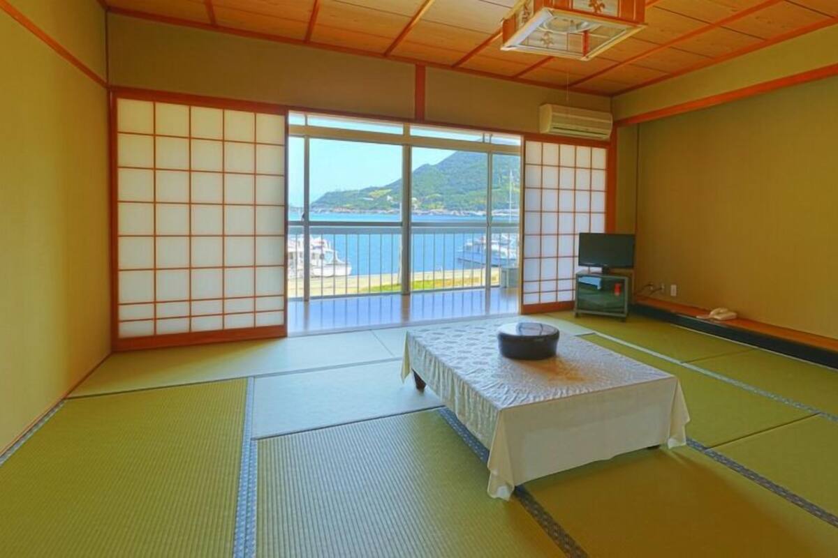 Ein traditioneller japanischer Raum mit Tatami-Bodenbelag, einem Tatami-Tisch, einem Futonbett, einem Fernseher und Blick auf das Meer.