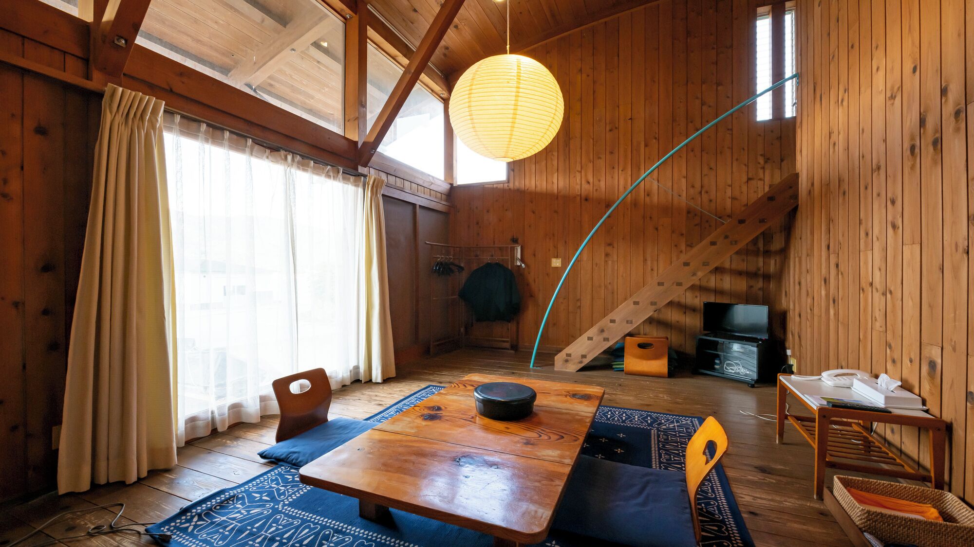 A wooden interior with a dining table, chairs, a hanging light, and a staircase.