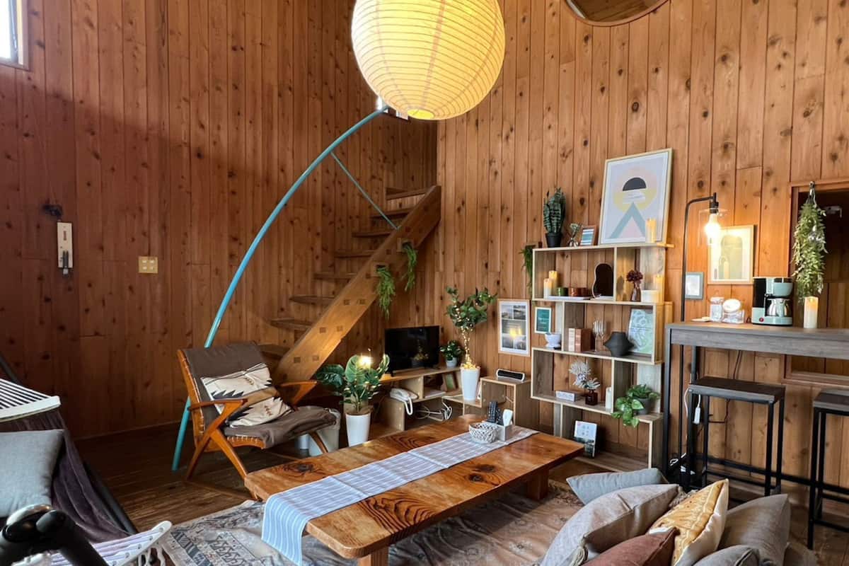Ein gemütliches Zimmer mit Holzboden, einer Holzbank, einem Tisch mit gestreiftem Tischläufer, einem Fernseher und einem Bücherregal mit diversen Gegenständen.