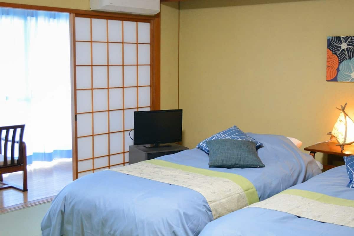 Ein Hotelzimmer mit zwei Betten, einem Fernseher, einem Schreibtisch, einem Sessel und einem Bild an der Wand.