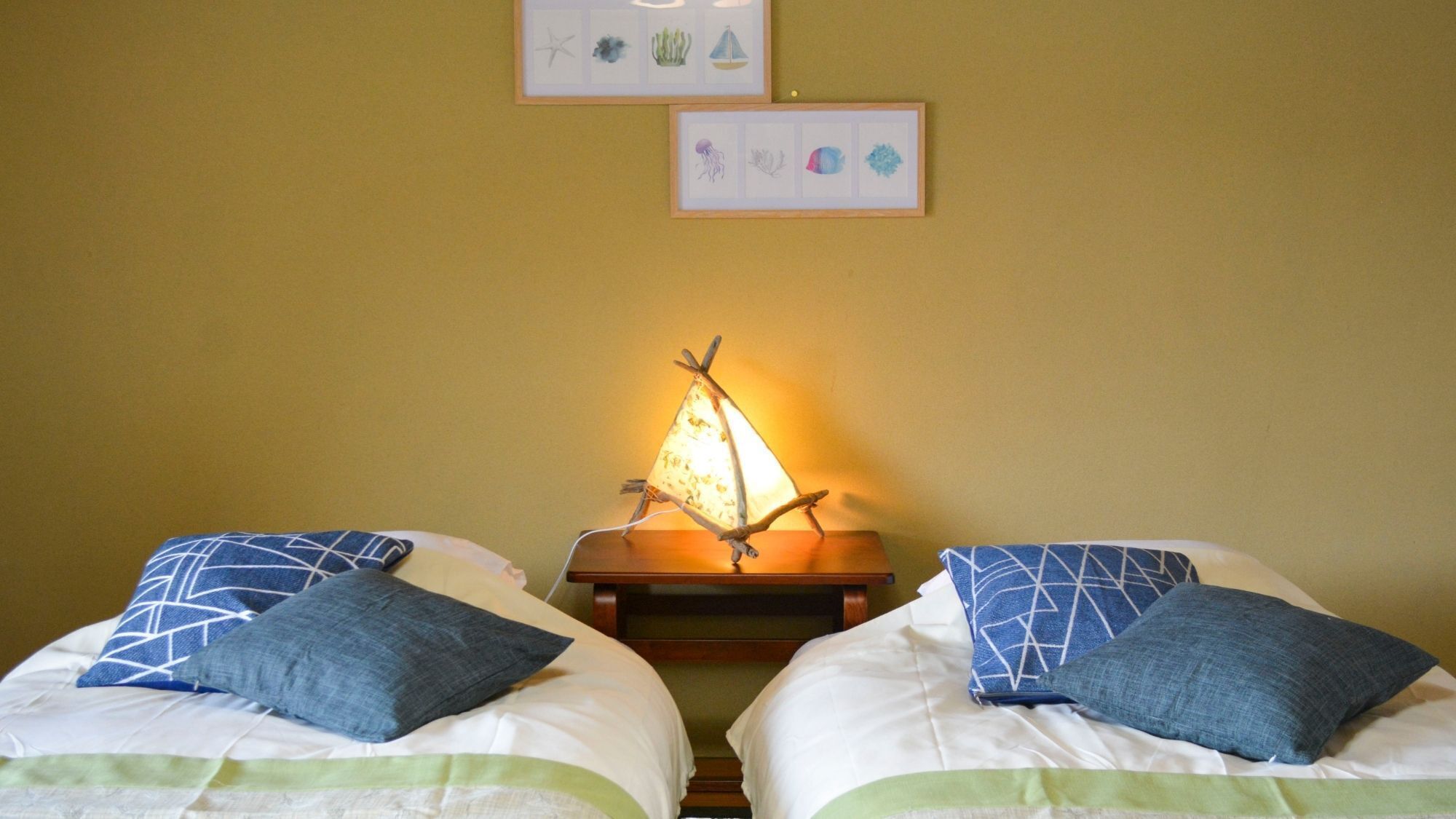Deux lits avec des oreillers bleus, une table de chevet avec une lampe et deux tableaux encadrés au mur.