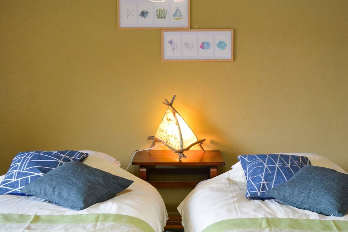 Zwei Betten mit blauen Kissen, ein Nachttisch mit einer Lampe und zwei gerahmte Bilder an der Wand.