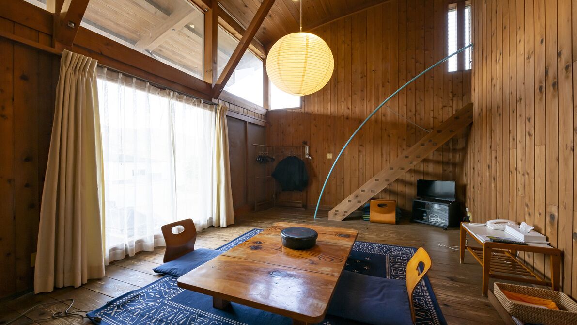 A wooden interior with a dining table, chairs, a hanging light fixture, and a staircase.