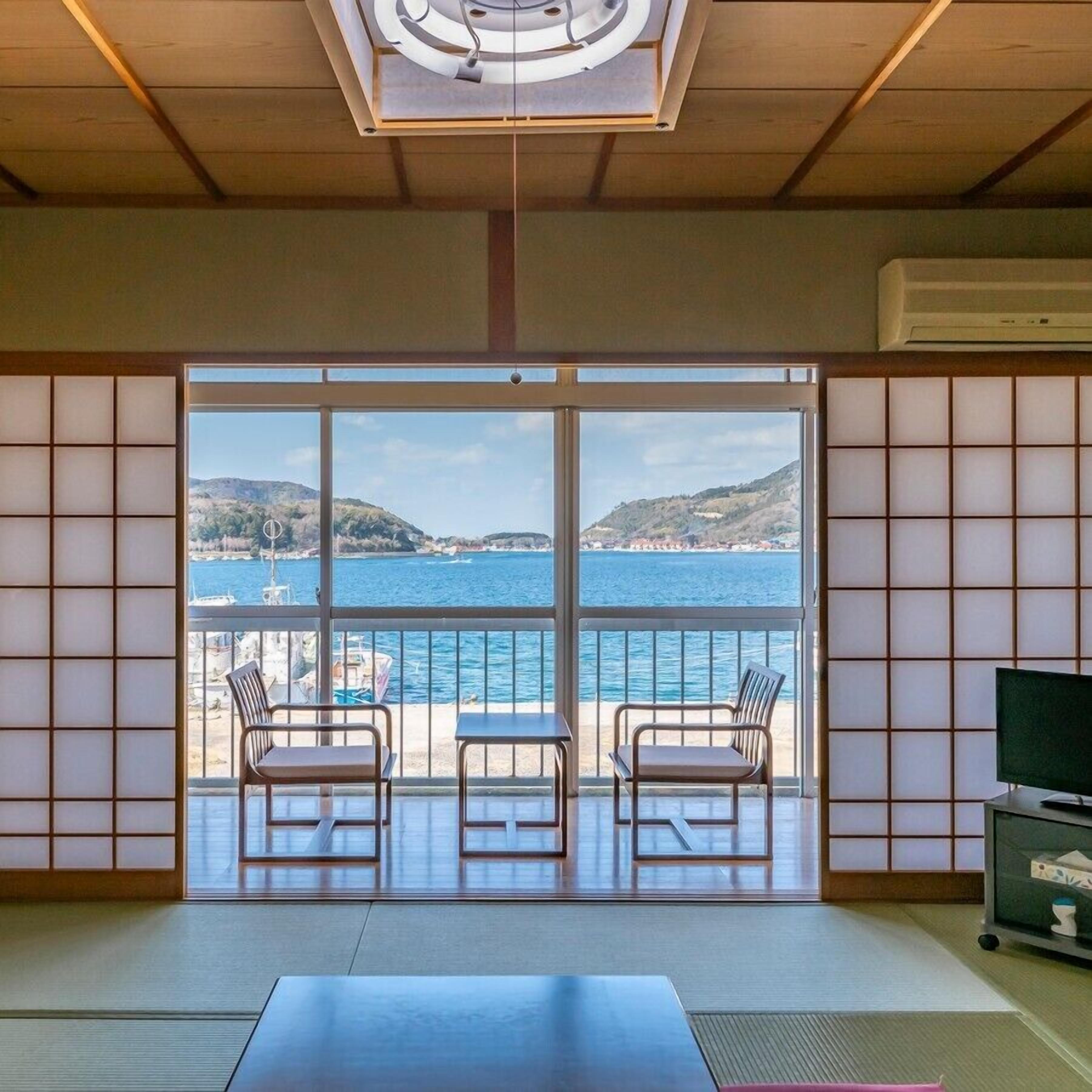 Une pièce dotée d’un sol recouvert de tatamis, une porte-fenêtre coulissante donnant sur un balcon avec vue sur la mer et les collines, et un téléviseur posé sur un meuble.
