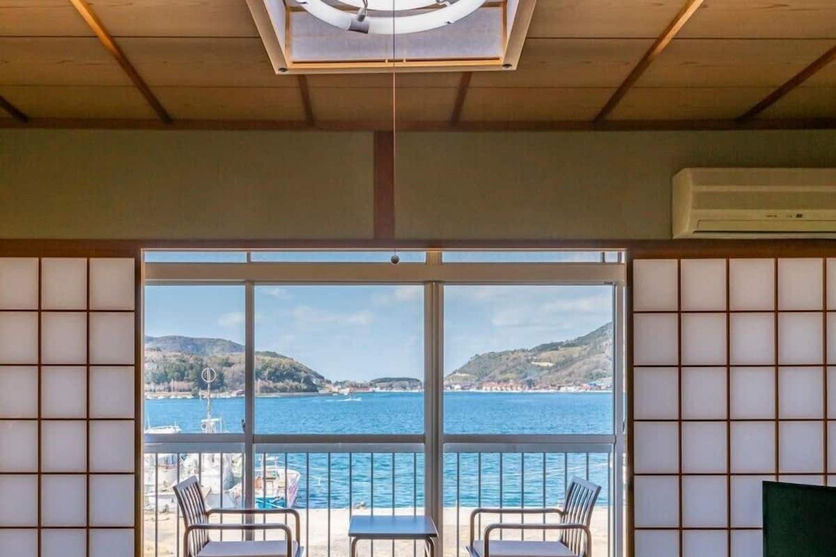 Ein Zimmer mit Tatami-Boden, einer Schiebetür zu einem Balkon mit Blick auf Meer und Hügel und einem Fernseher auf einem Podest.