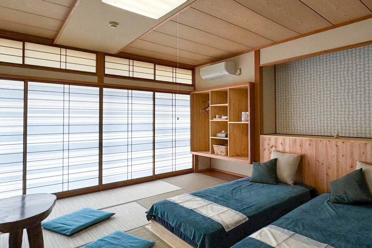 Ein traditioneller japanischer Raum mit Tatami-Boden, Futonbett, integriertem Ablagefach, Fernseher und großem Fenster mit Shoji-Schirmen.