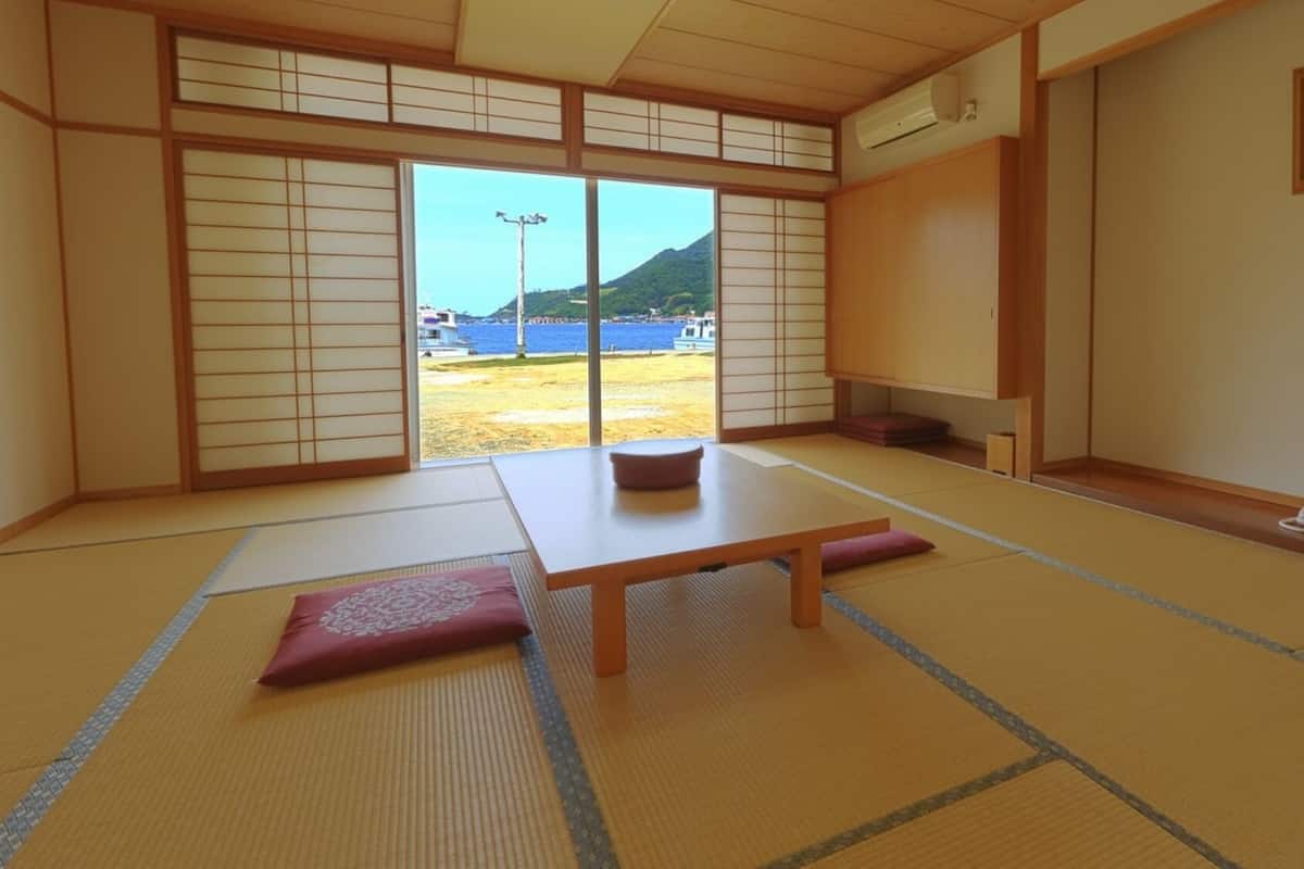 Ein Zimmer mit Tatami-Matten, einer Schiebetür zu einem Balkon, einem niedrigen Tisch und einem Kissen.