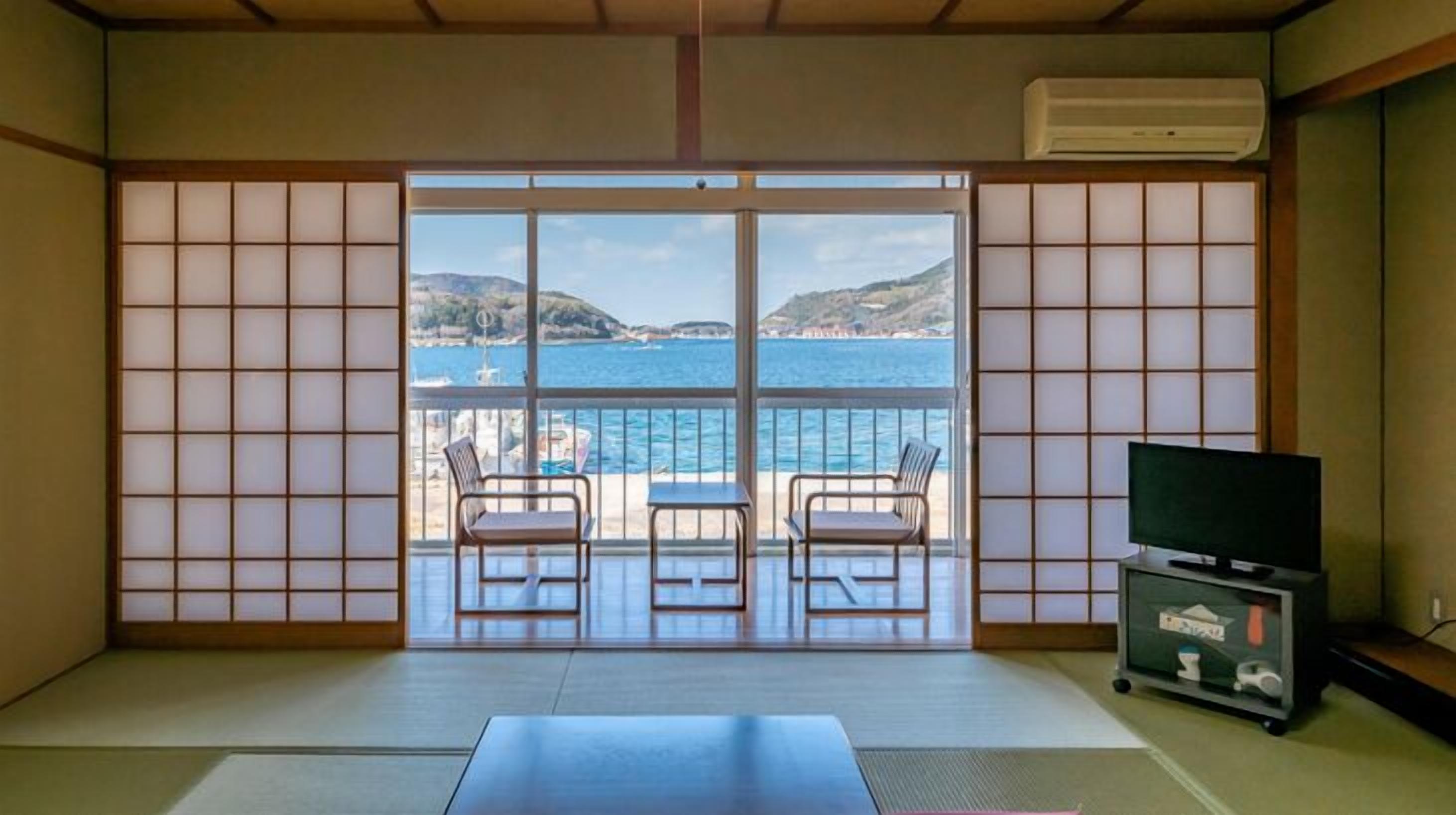 Une pièce dotée d’un sol recouvert de tatamis, une porte-fenêtre coulissante donnant sur un balcon avec vue sur la mer et les collines, deux fauteuils et un meuble pour la télévision avec une télécommande.