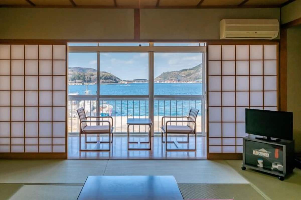 Ein Zimmer mit Tatami-Boden, einer Glastür, die auf einen Balkon mit Blick auf das Meer und die Hügel führt, zwei Stühle und eine TV-Konsole mit Fernbedienung.