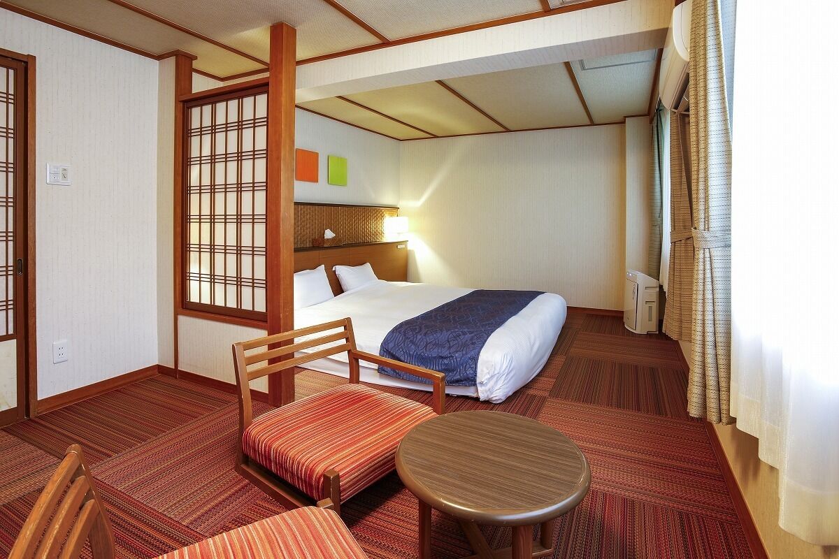 A hotel room with a bed, a chair, a small table, a window with curtains, and a wall with a decorative screen.