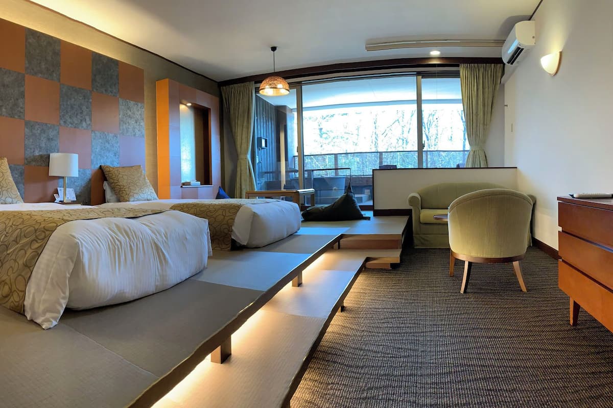 Habitación de hotel con dos camas, un escritorio, una silla y un amplio ventanal con cortinas.